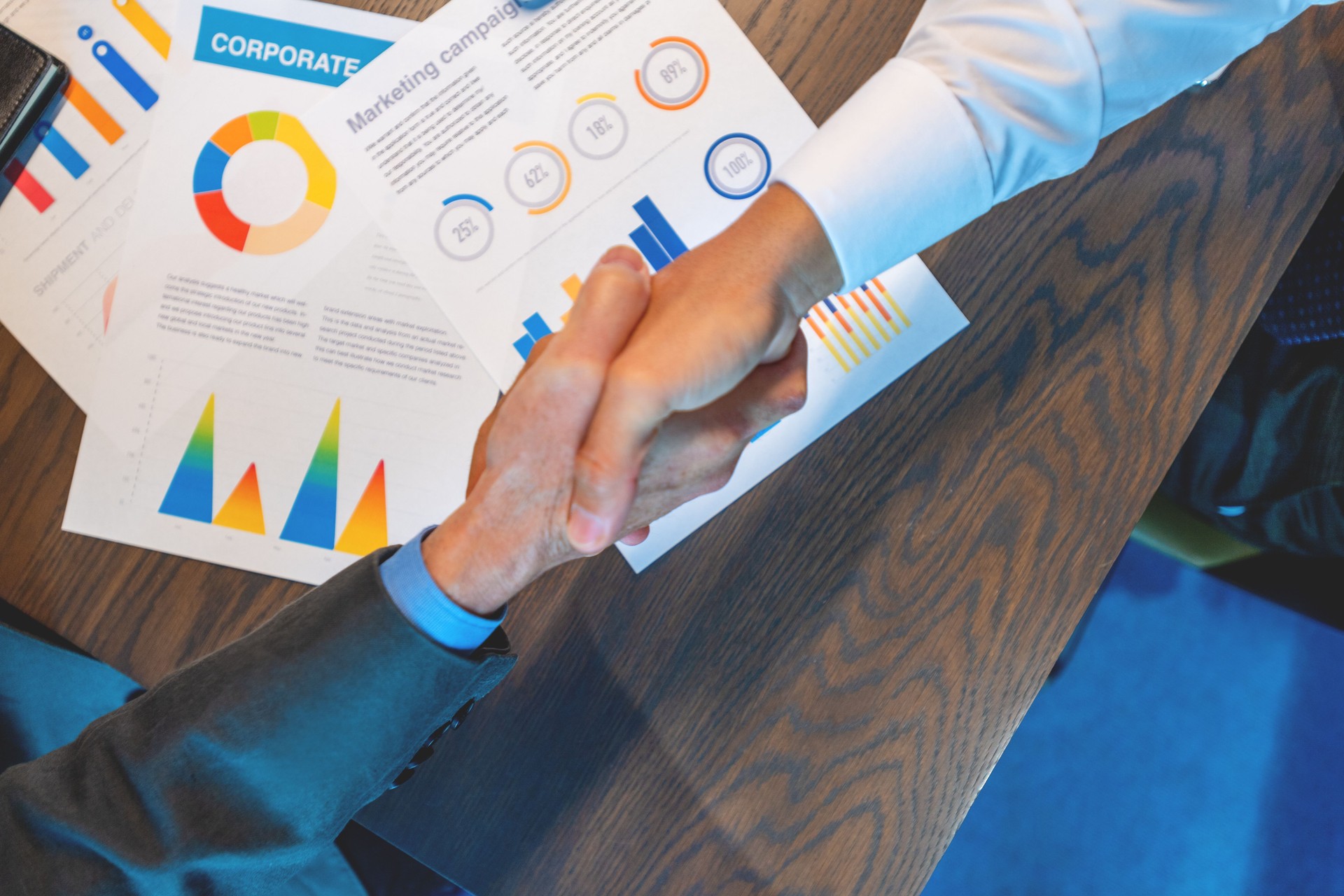 Businessmen shaking hands. Both are dressed in business clothing and there is paperwork with finance graphs and charts